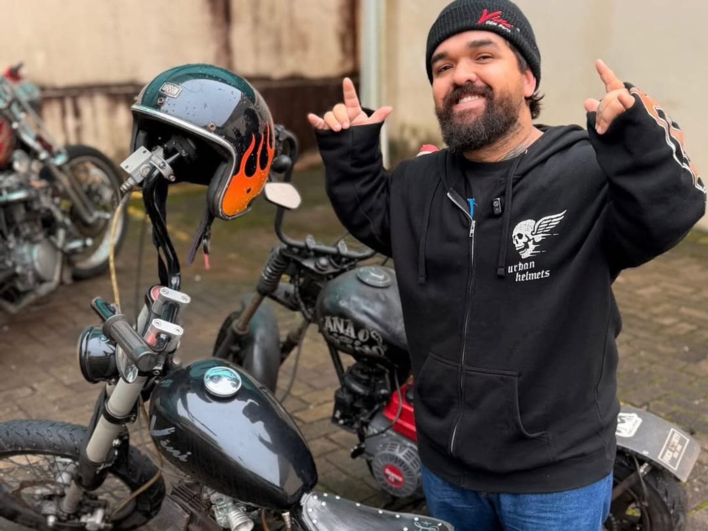Boladão ao lado de motos custom fazendo sinal de rock com as duas mãos